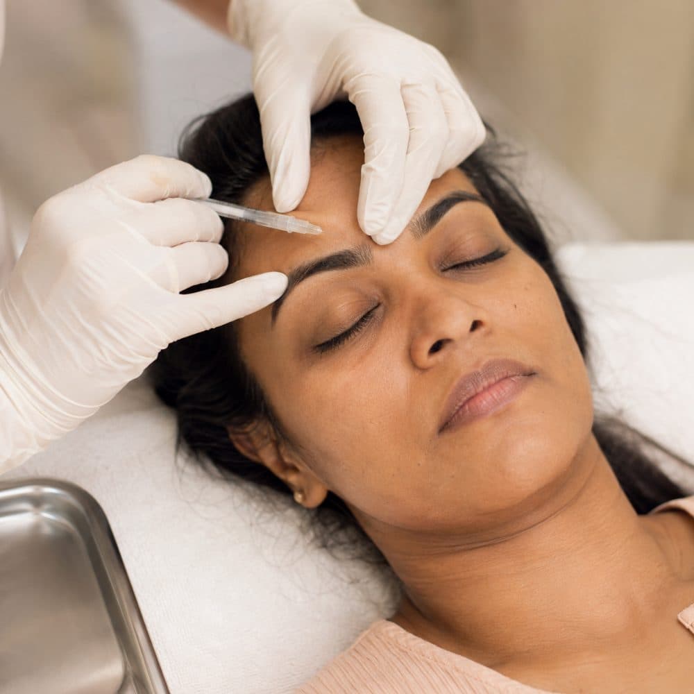 Gloved hands administering a fine injection to a patient's brow.